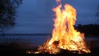 Ein großes, helles Feuer brennt am Ufer eines Sees bei Dämmerung. Die Flammen lodern hoch und erzeugen ein warmes Licht in der dunklen Umgebung. Im Hintergrund sind schemenhaft Bäume und Wasser zu erkennen.