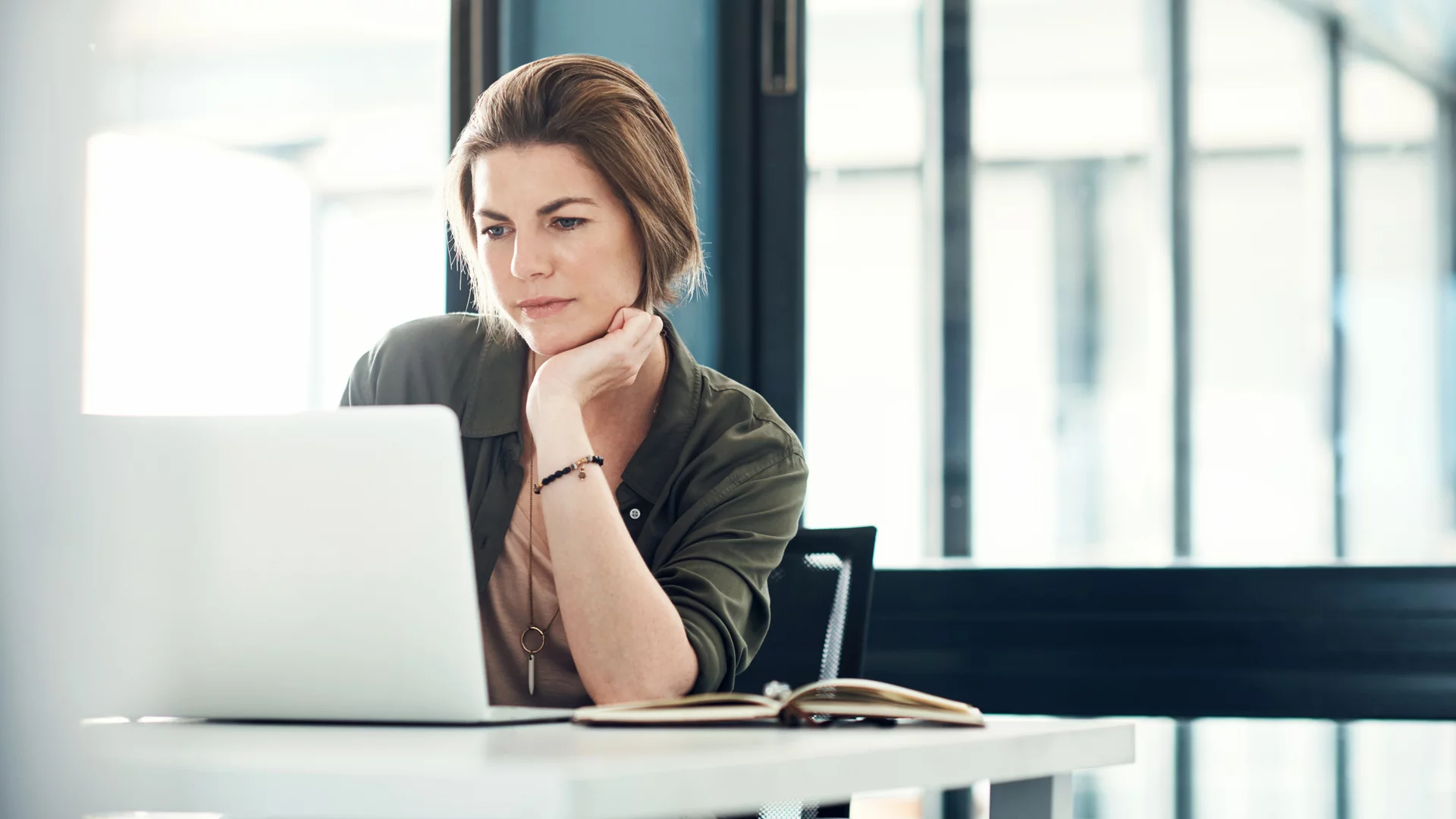 Eine nachdenkliche Frau sitzt an einem Tisch und schaut auf einen Laptop. Sie hat die Hand am Kinn gestützt und wirkt konzentriert. Im Hintergrund sind große Fenster, die Tageslicht hereinlassen.