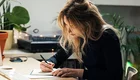 Eine Person mit langen, lockigen Haaren sitzt an einem Tisch und schreibt in ein Notizbuch. Umgeben von Pflanzen und einem Computer, konzentriert sie sich auf ihre Notizen. Der Raum wirkt freundlich und einladend.