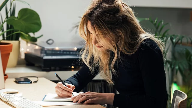 Eine Person mit langen, lockigen Haaren sitzt an einem Tisch und schreibt in ein Notizbuch. Umgeben von Pflanzen und einem Computer, konzentriert sie sich auf ihre Notizen. Der Raum wirkt freundlich und einladend.