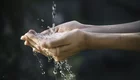Eine Person hält ihre offenen Hände unter einem Wasserstrom, der aus der Luft herunterfällt. Wasser spritzt und fließt durch die Hände, was ein Gefühl von Frische und Lebendigkeit vermittelt. Die Hände sind zart und verhältnismäßig nah an der Kamera.