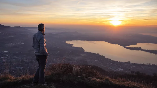 Ein Mann steht auf einem Hügel und blickt auf einen See, während die Sonne am Horizont untergeht. Die warmen Farben des Sonnenuntergangs spiegeln sich im Wasser und schaffen eine friedliche Atmosphäre.
