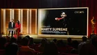 Die Szene zeigt eine Oscar-Verleihung mit zwei Moderatoren auf der Bühne. Im Hintergrund wird der Film "Marty Supreme" als bester Film nominiert, begleitet von Informationen über die Produzenten. Der Raum ist gefüllt mit applaudierenden Zuschauern.