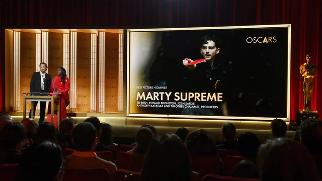 Die Szene zeigt eine Oscar-Verleihung mit zwei Moderatoren auf der Bühne. Im Hintergrund wird der Film "Marty Supreme" als bester Film nominiert, begleitet von Informationen über die Produzenten. Der Raum ist gefüllt mit applaudierenden Zuschauern.