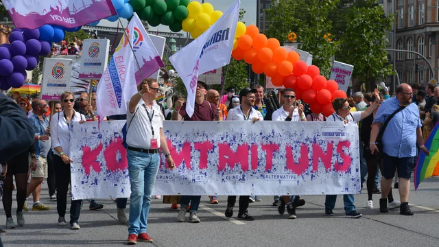 Eine Gruppe von Menschen nimmt an einer Parade teil und hält ein großes Banner mit der Aufschrift „KOMMT MIT UNS“. Sie tragen verschiedene Fahnen und stehen unter einem bunten Luftballonbogen. Die Atmosphäre ist festlich und die Teilnehmer wirken fröhlich und engagiert.