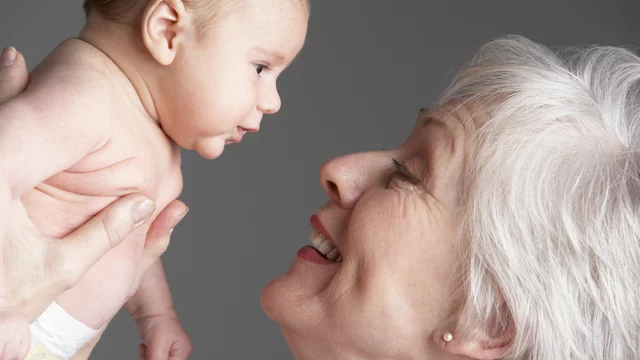 Eine ältere Frau hält ein Baby in den Händen, während sie liebevoll in dessen Gesicht schaut. Beide lächeln, was eine herzliche Verbindung zwischen den zwei Generationen ausdrückt. Der Hintergrund ist neutral und unterstützt den Fokus auf die beiden.