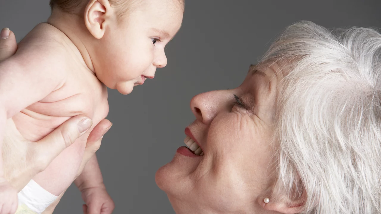 Eine ältere Frau hält ein Baby in den Händen, während sie liebevoll in dessen Gesicht schaut. Beide lächeln, was eine herzliche Verbindung zwischen den zwei Generationen ausdrückt. Der Hintergrund ist neutral und unterstützt den Fokus auf die beiden.