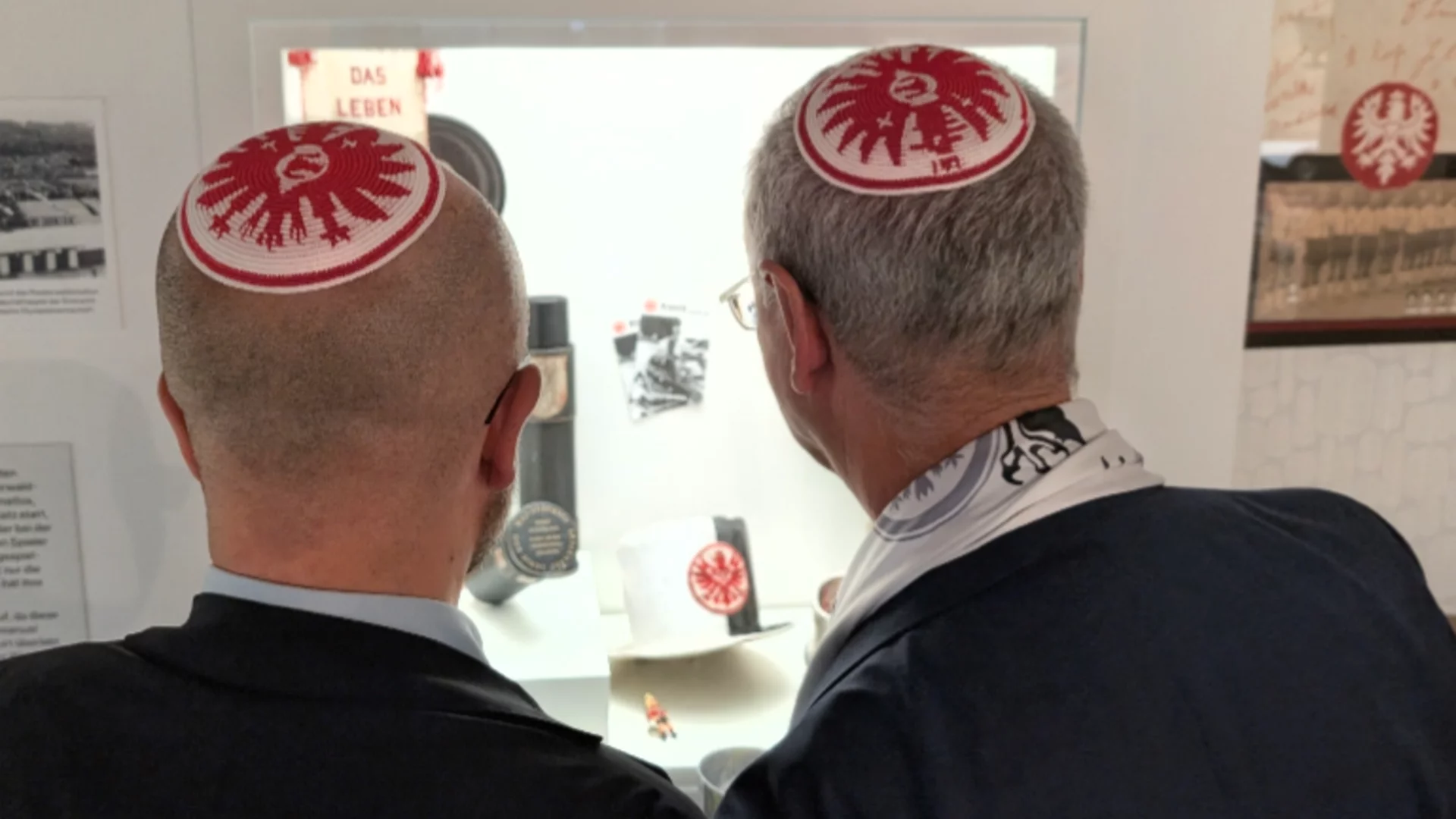 Zwei Männer, eine Glatze und graues Haar, stehen nebeneinander und betrachten eine Vitrine in einem Museum. Beide tragen Kippa, die mit dem Logo von Eintracht Frankfurt verziert sind. Im Hintergrund sind Ausstellungsobjekte zu sehen, die mit den Themen Leben und Kultur verbunden sind.
