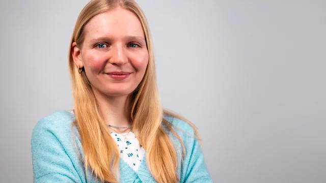 Eine junge Frau mit langen, blonden Haaren lächelt freundlich. Sie trägt einen hellblauen Pullover über einem weißen Oberteil mit einem Muster. Die Frau hat die Arme verschränkt und steht vor einem neutralen Hintergrund.