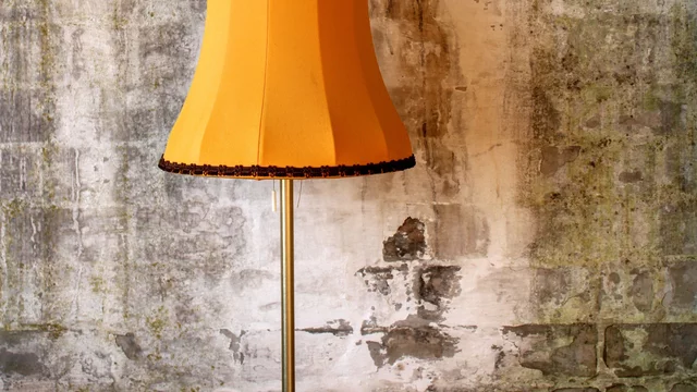 Eine orangefarbene Stehlampe mit einer geschwungenen Schirmform steht vor einer rauen, grauen Wand. Die Lampe hat einen metallischen Standfuß und einen dekorativen Rand. Der Gesamteindruck vermittelt eine vintage oder retro Ästhetik.