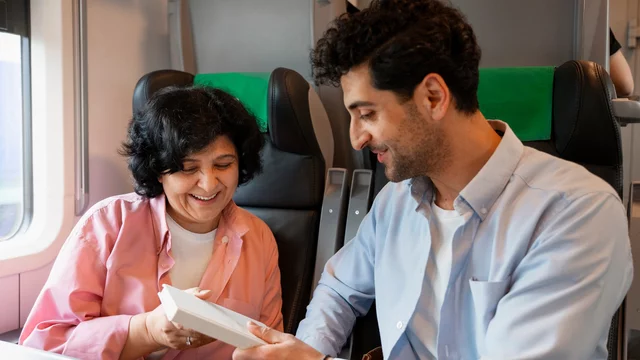 Auf dem Bild sind ein Mann und eine Frau in einem Zug zu sehen. Die Frau lächelt und hält ein Buch in der Hand, während der Mann ihr aufmerksam zuschaut. Die beiden scheinen sich über das Buch zu freuen.