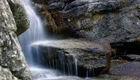 Ein kleiner Wasserfall fließt sanft über glatte, graue Felsen. Das Wasser sprudelt und erzeugt ein beruhigendes Geräusch, während es in einen klaren, ruhigen Bereich darunter rinnt. Umgebende Steine und etwas trockenes Gras rahmen die Szene ein.