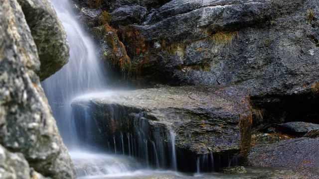 Ein kleiner Wasserfall fließt sanft über glatte, graue Felsen. Das Wasser sprudelt und erzeugt ein beruhigendes Geräusch, während es in einen klaren, ruhigen Bereich darunter rinnt. Umgebende Steine und etwas trockenes Gras rahmen die Szene ein.