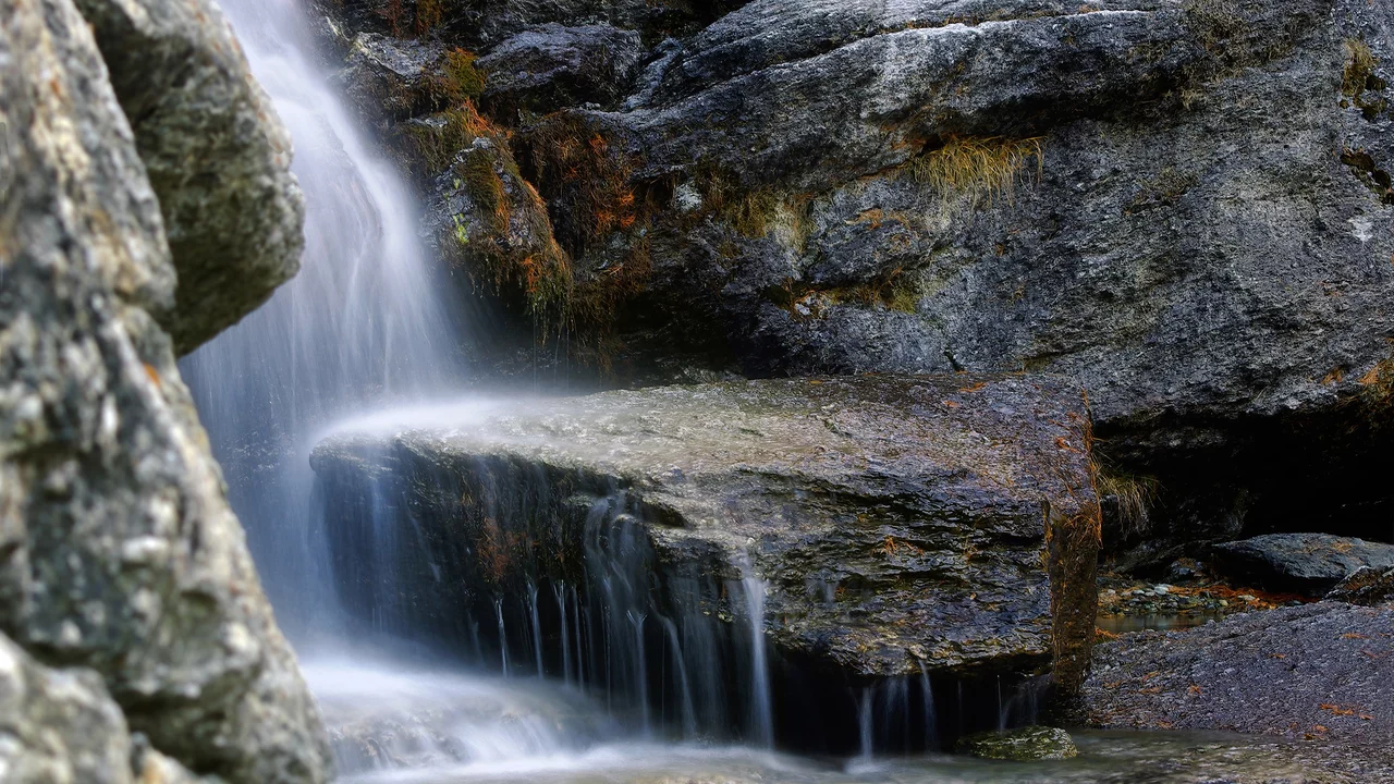 Ein kleiner Wasserfall fließt sanft über glatte, graue Felsen. Das Wasser sprudelt und erzeugt ein beruhigendes Geräusch, während es in einen klaren, ruhigen Bereich darunter rinnt. Umgebende Steine und etwas trockenes Gras rahmen die Szene ein.