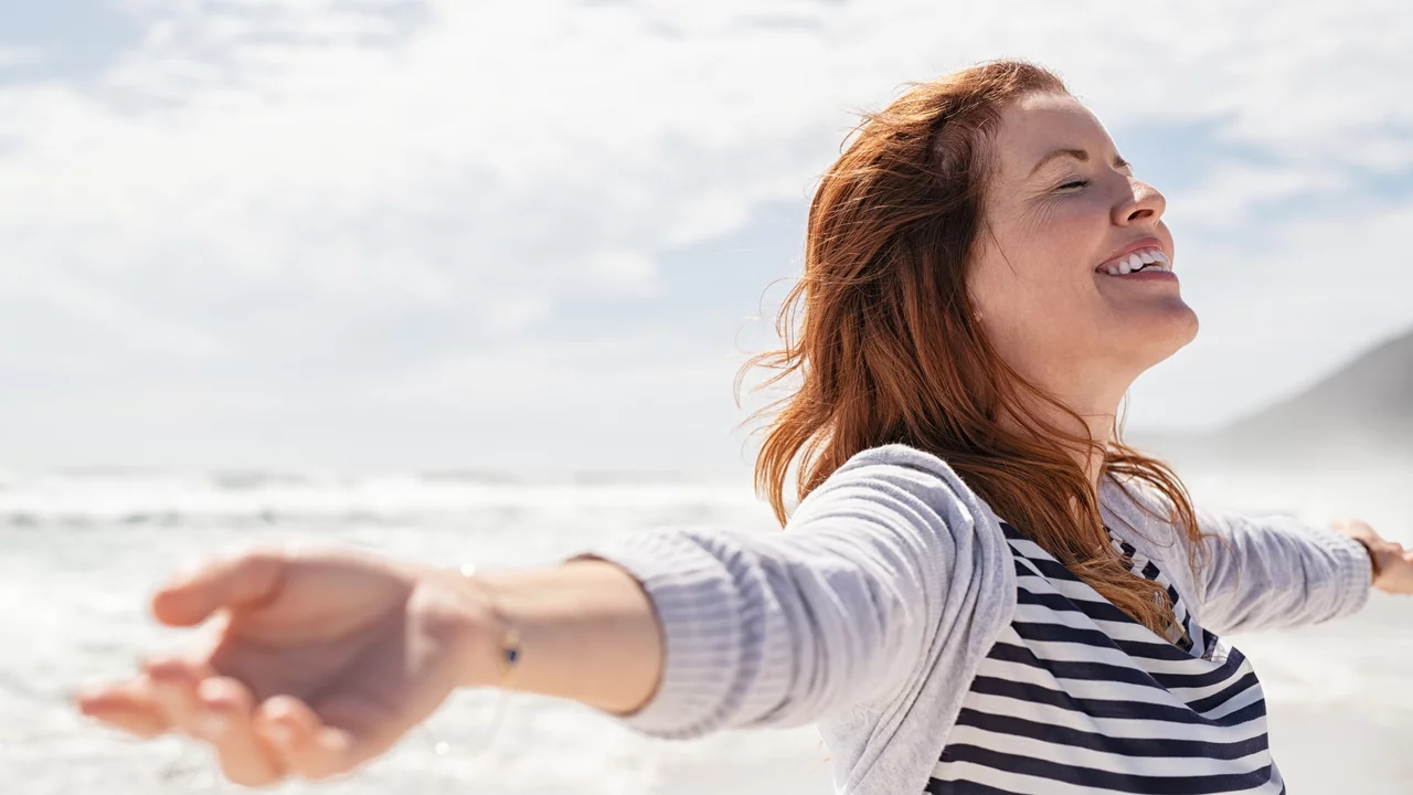 Eine lächelnde Frau steht am Strand mit weit geöffneten Armen. Sie genießt den Moment und scheint sich über die frische Luft und die Nähe zum Meer zu freuen. Ihr Haar weht im Wind, und die Atmosphäre wirkt entspannt und freundlich.