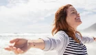 Eine lächelnde Frau steht am Strand mit weit geöffneten Armen. Sie genießt den Moment und scheint sich über die frische Luft und die Nähe zum Meer zu freuen. Ihr Haar weht im Wind, und die Atmosphäre wirkt entspannt und freundlich.