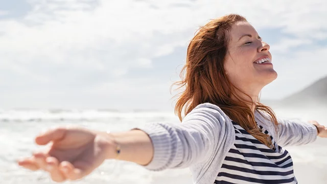 Eine lächelnde Frau steht am Strand mit weit geöffneten Armen. Sie genießt den Moment und scheint sich über die frische Luft und die Nähe zum Meer zu freuen. Ihr Haar weht im Wind, und die Atmosphäre wirkt entspannt und freundlich.