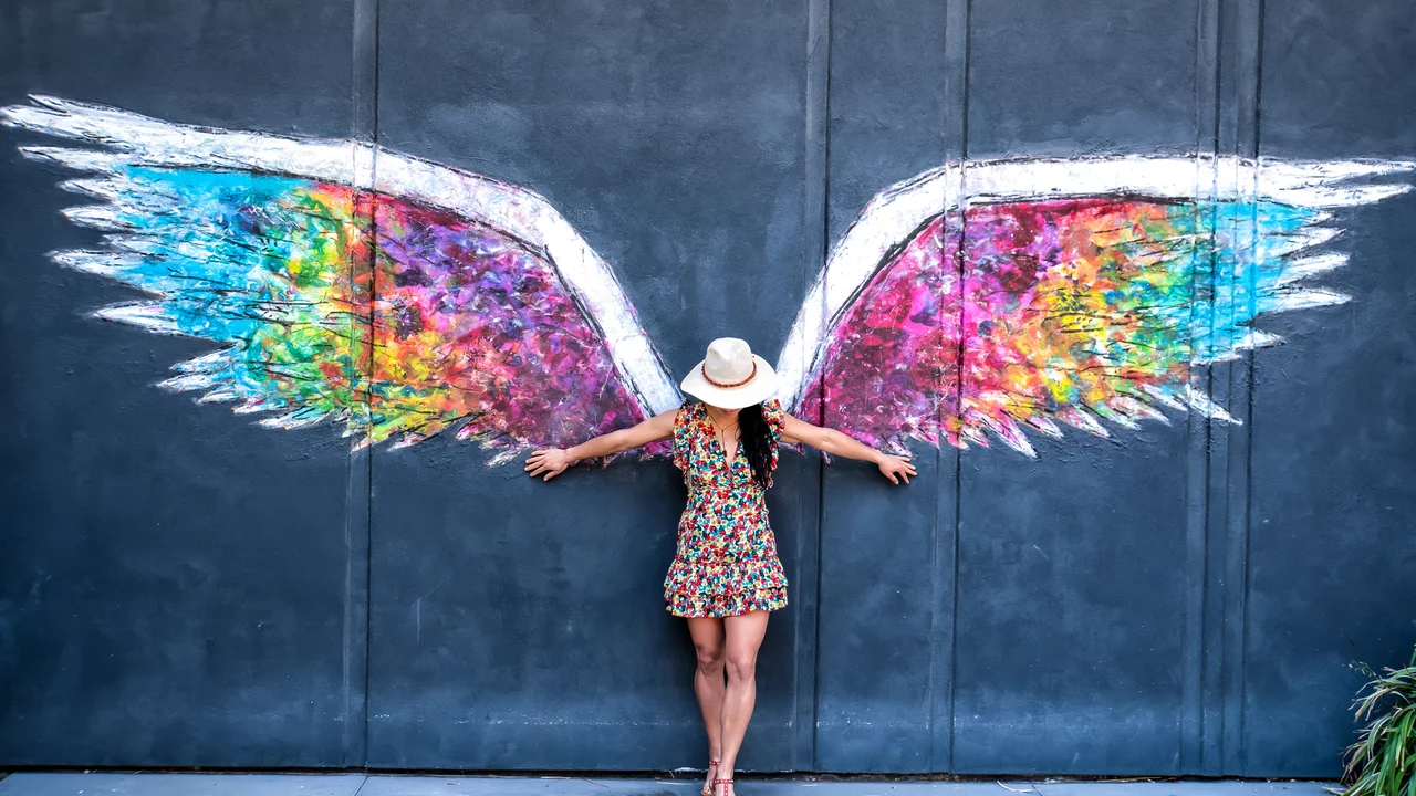 Eine Frau in einem bunten Kleid steht vor einer Wand mit farbenfrohen Flügeln. Sie breitet die Arme aus und berührt die Flügel, die in verschiedenen Farben leuchten. Ihr Hut und die lebhaften Farben erzeugen eine fröhliche Atmosphäre.
