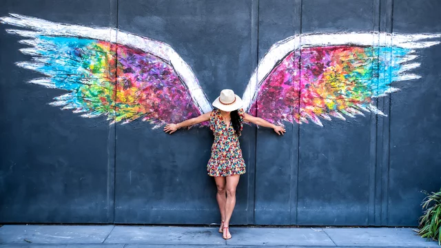 Eine Frau in einem bunten Kleid steht vor einer Wand mit farbenfrohen Flügeln. Sie breitet die Arme aus und berührt die Flügel, die in verschiedenen Farben leuchten. Ihr Hut und die lebhaften Farben erzeugen eine fröhliche Atmosphäre.