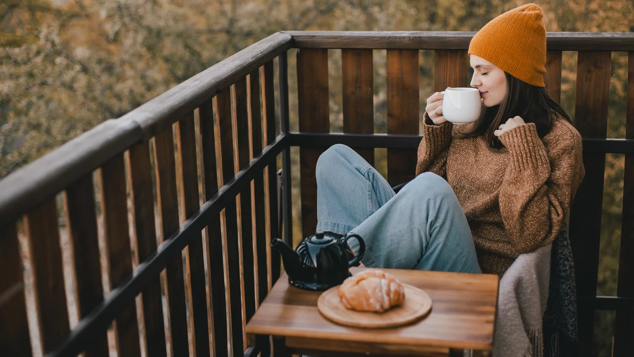Eine Frau sitzt auf einem Balkon, trägt einen orangefarbenen Hut und einen gemütlichen Pullover. Sie hält eine Tasse in der Hand und lächelt. Vor ihr befindet sich ein Tisch mit einem kleinen Kännchen und einem Croissant. Die Umgebung zeigt herbstliche Bäume.