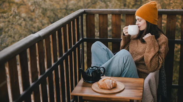 Eine Frau sitzt auf einem Balkon, trägt einen orangefarbenen Hut und einen gemütlichen Pullover. Sie hält eine Tasse in der Hand und lächelt. Vor ihr befindet sich ein Tisch mit einem kleinen Kännchen und einem Croissant. Die Umgebung zeigt herbstliche Bäume.