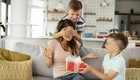 Eine Mutter sitzt auf einem Sofa und wird von ihrem älteren Sohn überrascht, der ihr die Augen bedeckt. Der jüngere Sohn hält ein Geschenk in den Händen und lächelt. Die Atmosphäre ist fröhlich und voller Überraschung.