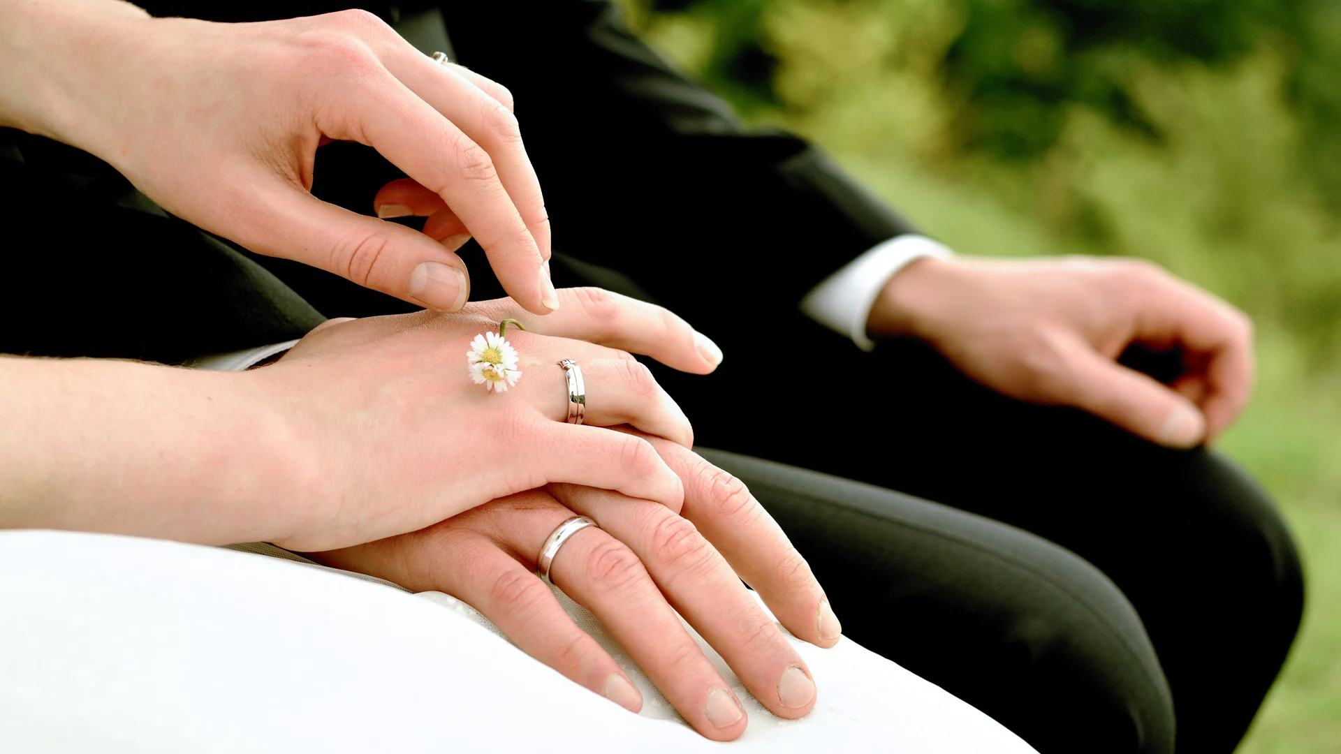 Hochzeitsring wird an der Hand des Bräutigams angebracht. Im Hintergrund sind zwei Hände zu sehen, eine trägt einen Ring, die andere ist sanft darauf gelegt. Der Fokus liegt auf der Symbolik der Verbundenheit und des Eheversprechens.
