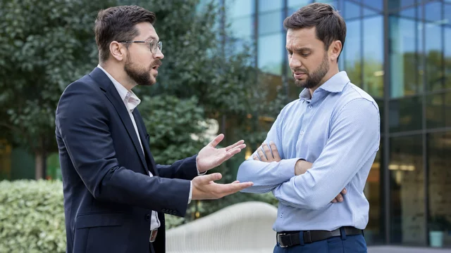 Zwei Männer stehen im Freien vor einem modernen Gebäude. Der Mann links, in einem schwarzen Anzug und mit Brille, spricht leidenschaftlich und gestikuliert. Der Mann rechts, im hellblauen Hemd, schaut nachdenklich und hat die Arme verschränkt. Es scheint eine angespannte Diskussion zu sein.