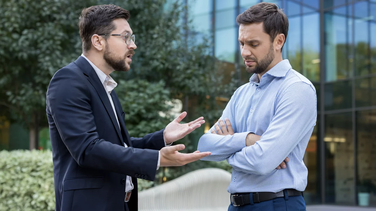 Zwei Männer stehen im Freien vor einem modernen Gebäude. Der Mann links, in einem schwarzen Anzug und mit Brille, spricht leidenschaftlich und gestikuliert. Der Mann rechts, im hellblauen Hemd, schaut nachdenklich und hat die Arme verschränkt. Es scheint eine angespannte Diskussion zu sein.