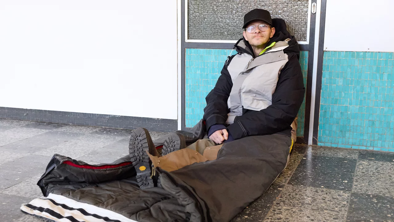 Eine Person ohne Obdach sitzt in einem Schlafsack auf dem Boden in einem Innenraum. Die Person trägt warme Kleidung, einschließlich einer Jacke und Mütze, und hat eine Brille auf. Der Hintergrund zeigt eine Wand mit blauen Fliesen und eine Glastür.
