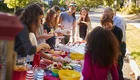 Eine Gruppe von Kindern und Erwachsenen steht um einen Tisch im Freien, der mit verschiedenen Speisen und Getränken gedeckt ist. Alle scheinen sich zu amüsieren und miteinander zu interagieren. Die Szene vermittelt eine freundliche, gesellige Atmosphäre.