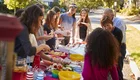Eine Gruppe von Kindern und Erwachsenen steht um einen Tisch im Freien, der mit verschiedenen Speisen und Getränken gedeckt ist. Alle scheinen sich zu amüsieren und miteinander zu interagieren. Die Szene vermittelt eine freundliche, gesellige Atmosphäre.
