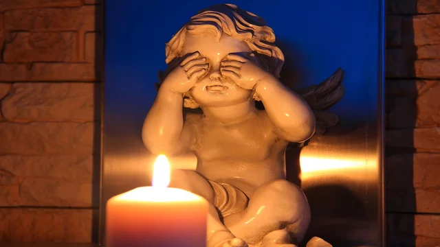 Eine kleine Statue eines Engels sitzt und hält sich die Augen zu. Vor ihr brennt eine Kerze, deren Licht eine warme Atmosphäre schafft. Die Statue drückt eine gefühlvolle, schützende Geste aus und vermittelt eine Botschaft von Trauer oder Nachdenklichkeit.