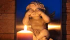 Eine kleine Statue eines Engels sitzt und hält sich die Augen zu. Vor ihr brennt eine Kerze, deren Licht eine warme Atmosphäre schafft. Die Statue drückt eine gefühlvolle, schützende Geste aus und vermittelt eine Botschaft von Trauer oder Nachdenklichkeit.