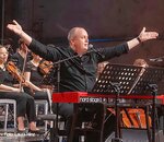 Ein Musiker sitzt an einem Keyboard und dirigiert mit beiden Armen eine Orchestergruppe im Hintergrund. Die Musikerinnen und Musiker sind in schwarzer Kleidung und konzentriert auf ihre Instrumente. Die Szene wirkt lebhaft und dynamisch.