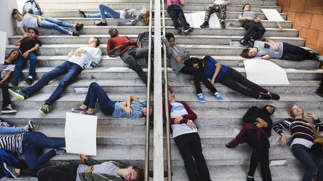 Gruppen von Jugendlichen liegen auf einer Treppe, einige halten leere Plakate. Die Szene vermittelt eine Botschaft des Protests oder der Aktivität, mit verschiedenen Haltungen und Ausdrücken der Teilnehmer.