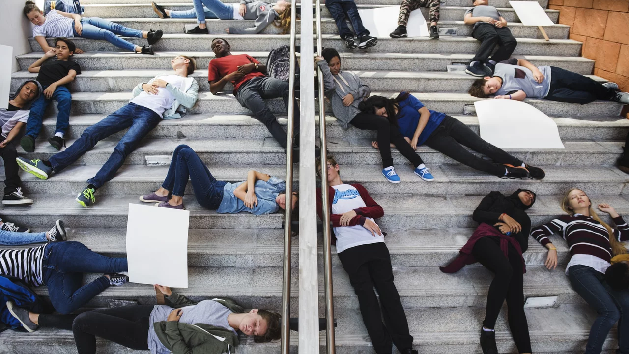 Gruppen von Jugendlichen liegen auf einer Treppe, einige halten leere Plakate. Die Szene vermittelt eine Botschaft des Protests oder der Aktivität, mit verschiedenen Haltungen und Ausdrücken der Teilnehmer.