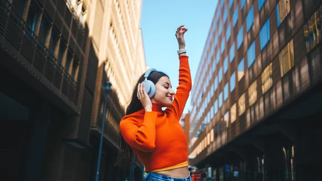 Eine junge Frau in einem orangefarbenen Pullover tanzt und hört Musik mit Kopfhörern. Sie hebt eine Hand in die Luft, während sie sich in einer urbanen Umgebung zwischen modernen Gebäuden befindet, und strahlt Freude aus.