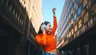 Eine junge Frau in einem orangefarbenen Pullover tanzt und hört Musik mit Kopfhörern. Sie hebt eine Hand in die Luft, während sie sich in einer urbanen Umgebung zwischen modernen Gebäuden befindet, und strahlt Freude aus.