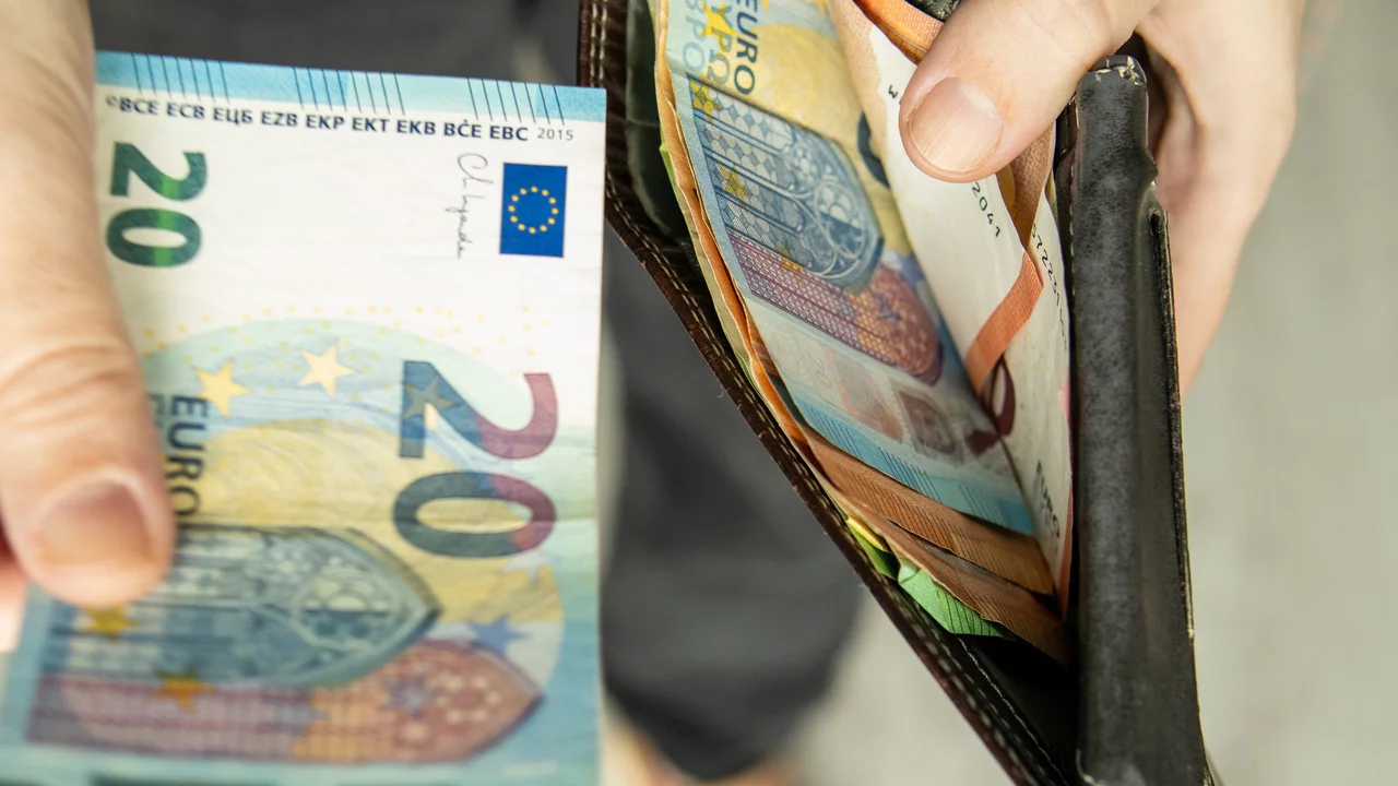 Eine Hand hält einen 20-Euro-Schein, während eine Geldbörse geöffnet ist und mehrere weitere Geldscheine sichtbar sind. Die Hand zeigt, dass der Schein entnommen wird, was auf eine finanzielle Transaktion hinweist.