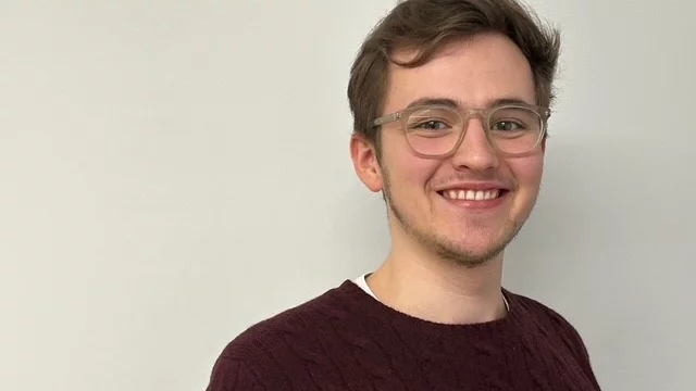 Ein junger Mann mit Brille lächelt in die Kamera. Er trägt einen dunkelroten, pulloverförmigen Sweater und steht vor einer hellen Wand. Sein freundlicher Gesichtsausdruck vermittelt eine positive Stimmung.