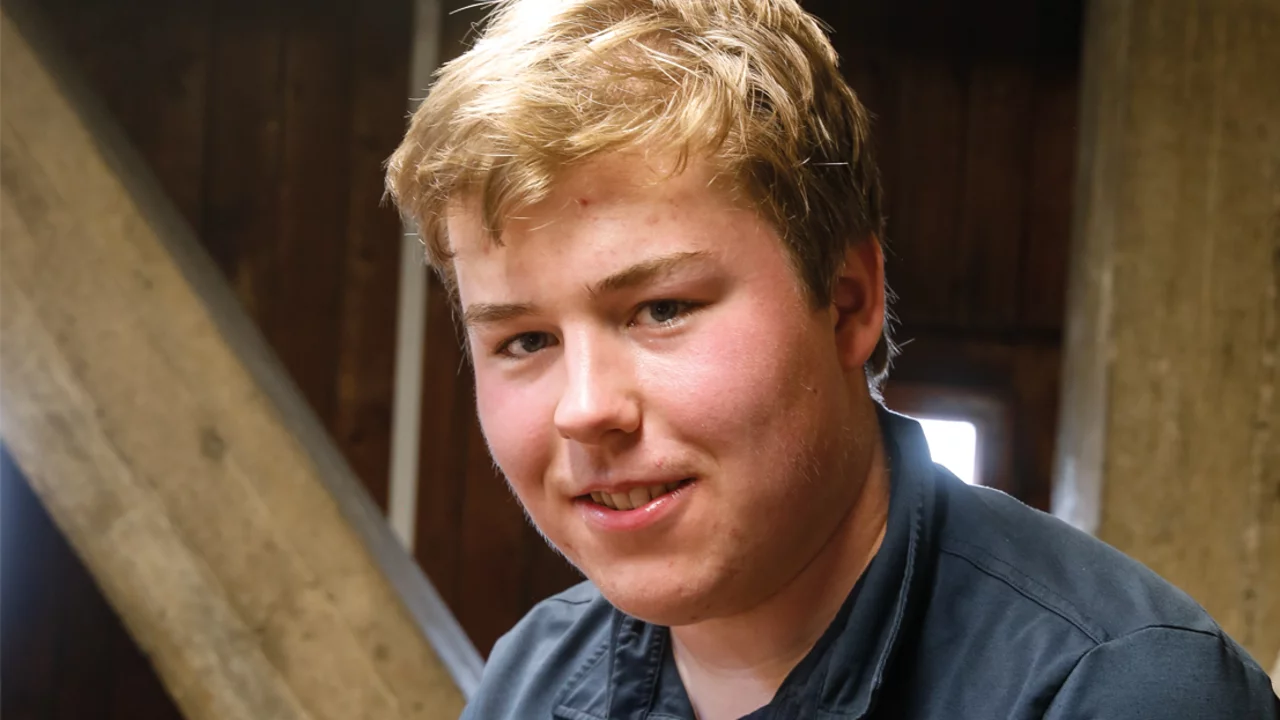 Ein junger Mann mit blonden Haaren lächelt in die Kamera. Er trägt ein dunkles Hemd und sitzt in einem Raum mit holzverkleideten Wänden. Im Hintergrund sind tragende Balken sichtbar.
