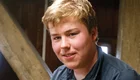 Ein junger Mann mit blonden Haaren lächelt in die Kamera. Er trägt ein dunkles Hemd und sitzt in einem Raum mit holzverkleideten Wänden. Im Hintergrund sind tragende Balken sichtbar.