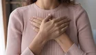 Eine lächelnde Frau steht mit verschränkten Händen vor der Brust da. Sie trägt einen rosa Pullover und hat lockige Haare. Der Hintergrund zeigt eine helle, gemütliche Innenraumgestaltung.
