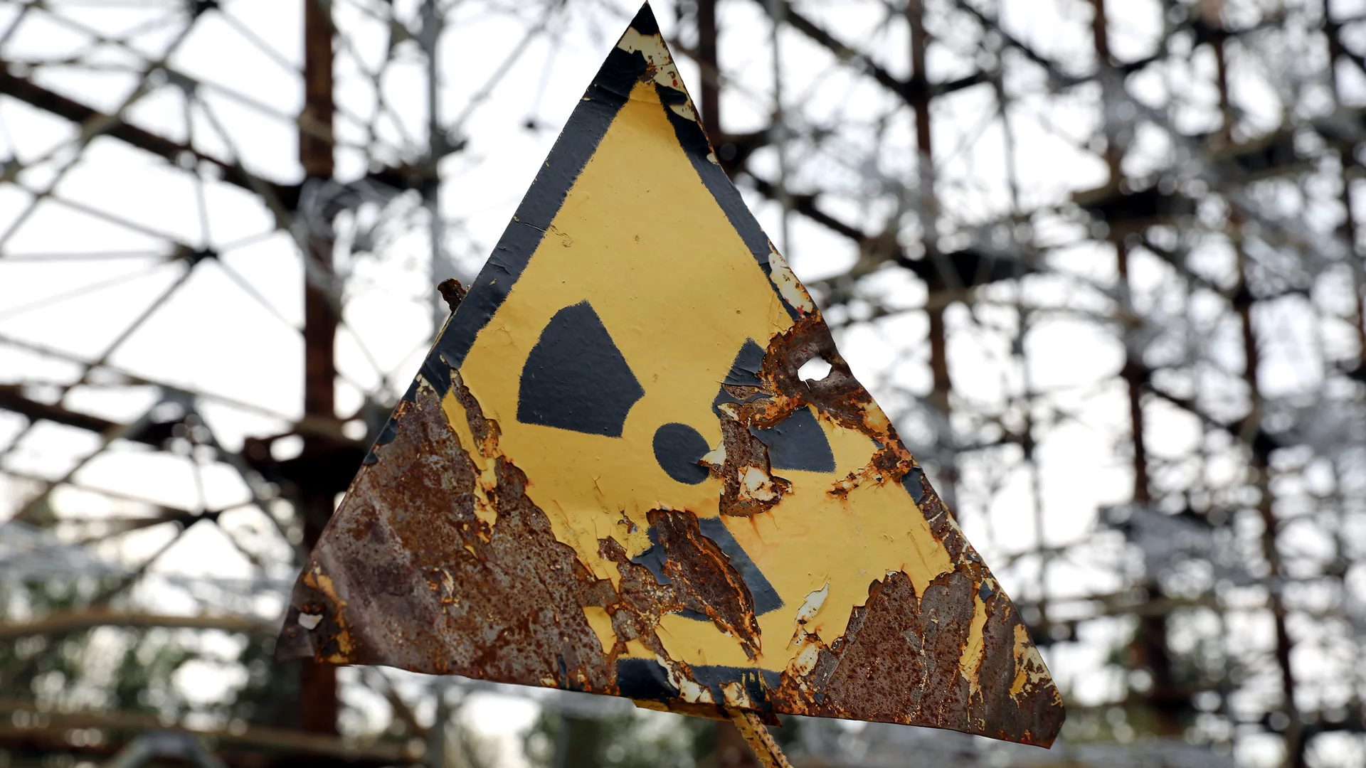 Ein abgenutztes, gelbes Warnschild in Form eines Dreiecks mit dem Symbol für radioaktive Strahlung. Der Hintergrund zeigt eine verfallene Struktur, die an eine industrielle Umgebung erinnert. Das Schild weist auf mögliche Gefahren hin.