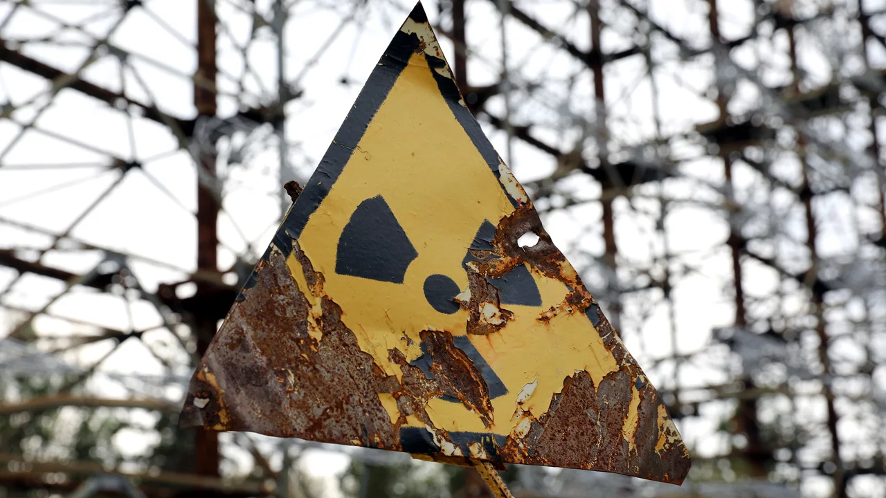 Ein abgenutztes, gelbes Warnschild in Form eines Dreiecks mit dem Symbol für radioaktive Strahlung. Der Hintergrund zeigt eine verfallene Struktur, die an eine industrielle Umgebung erinnert. Das Schild weist auf mögliche Gefahren hin.