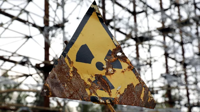 Ein abgenutztes, gelbes Warnschild in Form eines Dreiecks mit dem Symbol für radioaktive Strahlung. Der Hintergrund zeigt eine verfallene Struktur, die an eine industrielle Umgebung erinnert. Das Schild weist auf mögliche Gefahren hin.